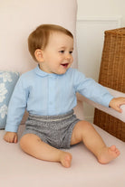 Smiling baby boy sitting on a pink armchair wearing a light blue shirt and gray tweed shorts