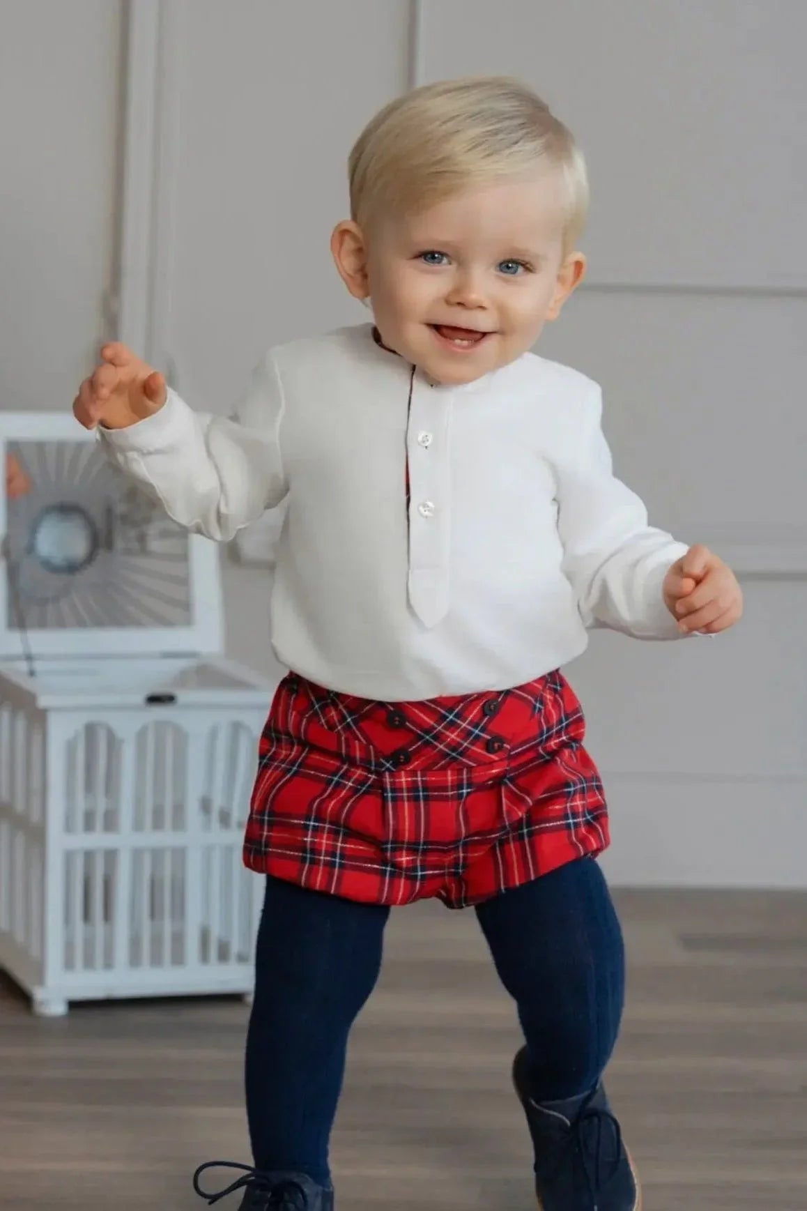 Smiling toddler in white buttoned shirt, red plaid shorts, navy tights, and navy shoes indoors