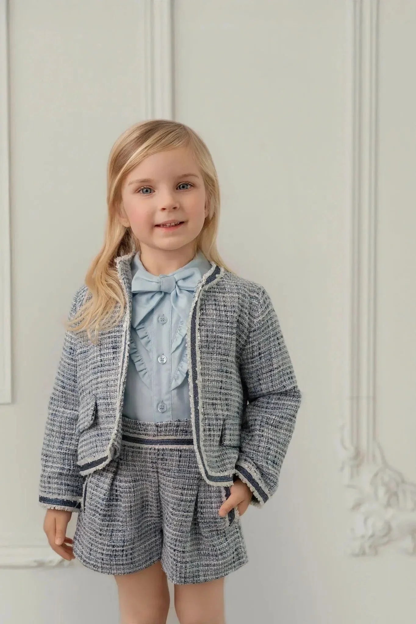 Young girl wearing blue tweed blazer, matching shorts, and light blue ruffled shirt with bow in elegant room