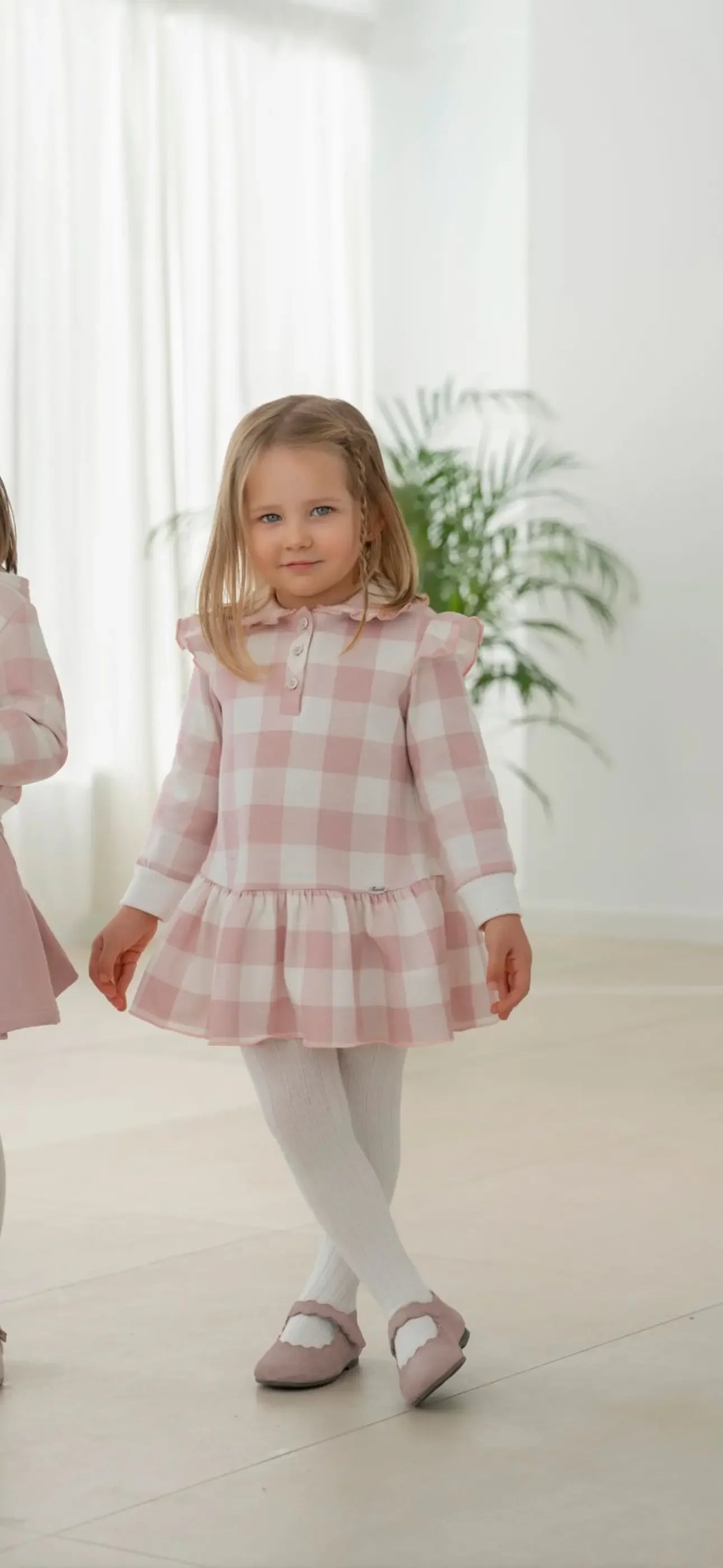 Young girl wearing pink and white check dress with white tights and pink shoes in bright room