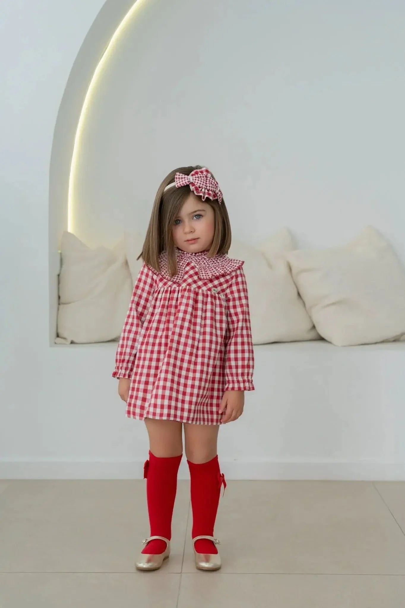 Little girl in red and white checkered dress with matching headband and red knee-high socks indoors