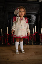 Toddler girl in a cream plaid dress with red ruffles and knee socks standing indoors by a fireplace with candles