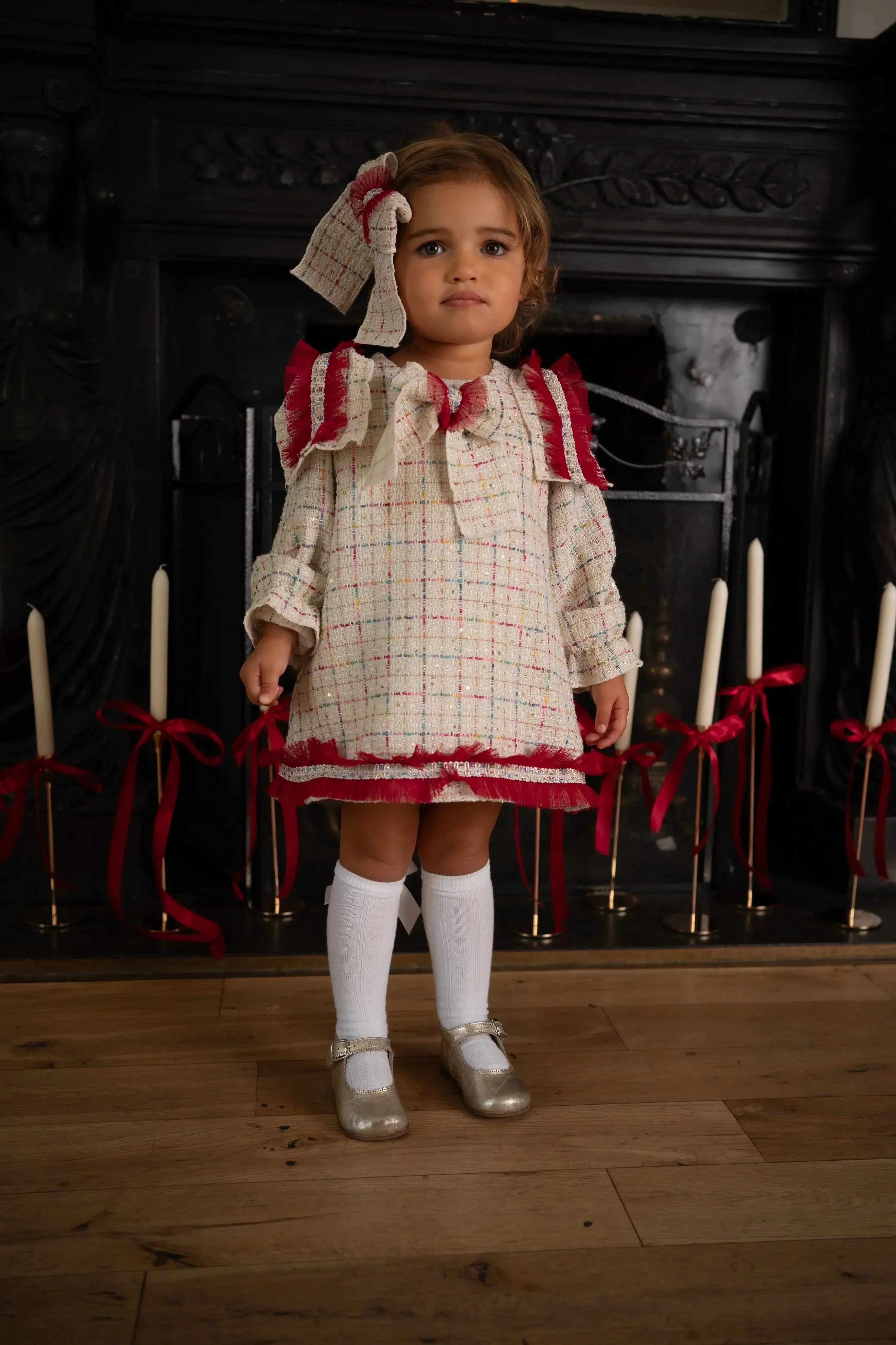 Toddler girl in a cream plaid dress with red ruffles and knee socks standing indoors by a fireplace with candles