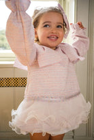 Smiling toddler girl in pink textured dress with ruffled tulle skirt and matching bow indoors