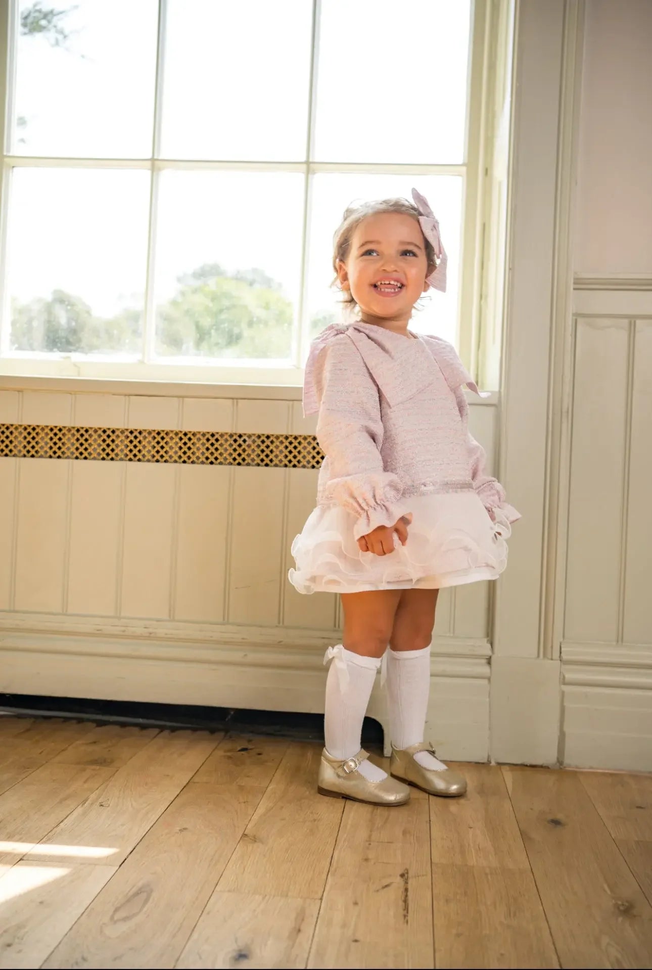 Smiling toddler girl in pink dress with ruffles, white knee-high socks, and gold shoes by window