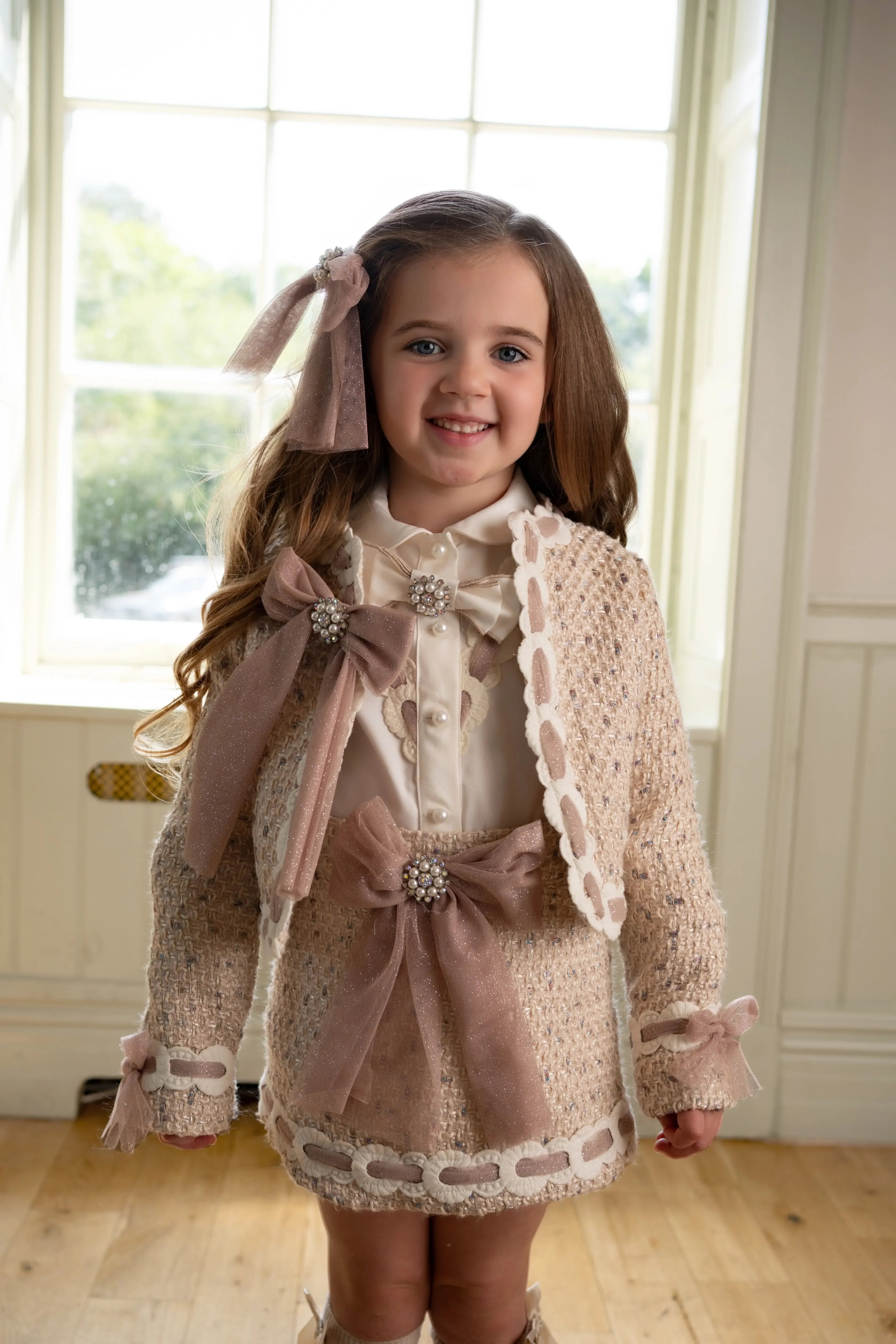 Smiling young girl in beige knit outfit with pearl bows, standing indoors near window