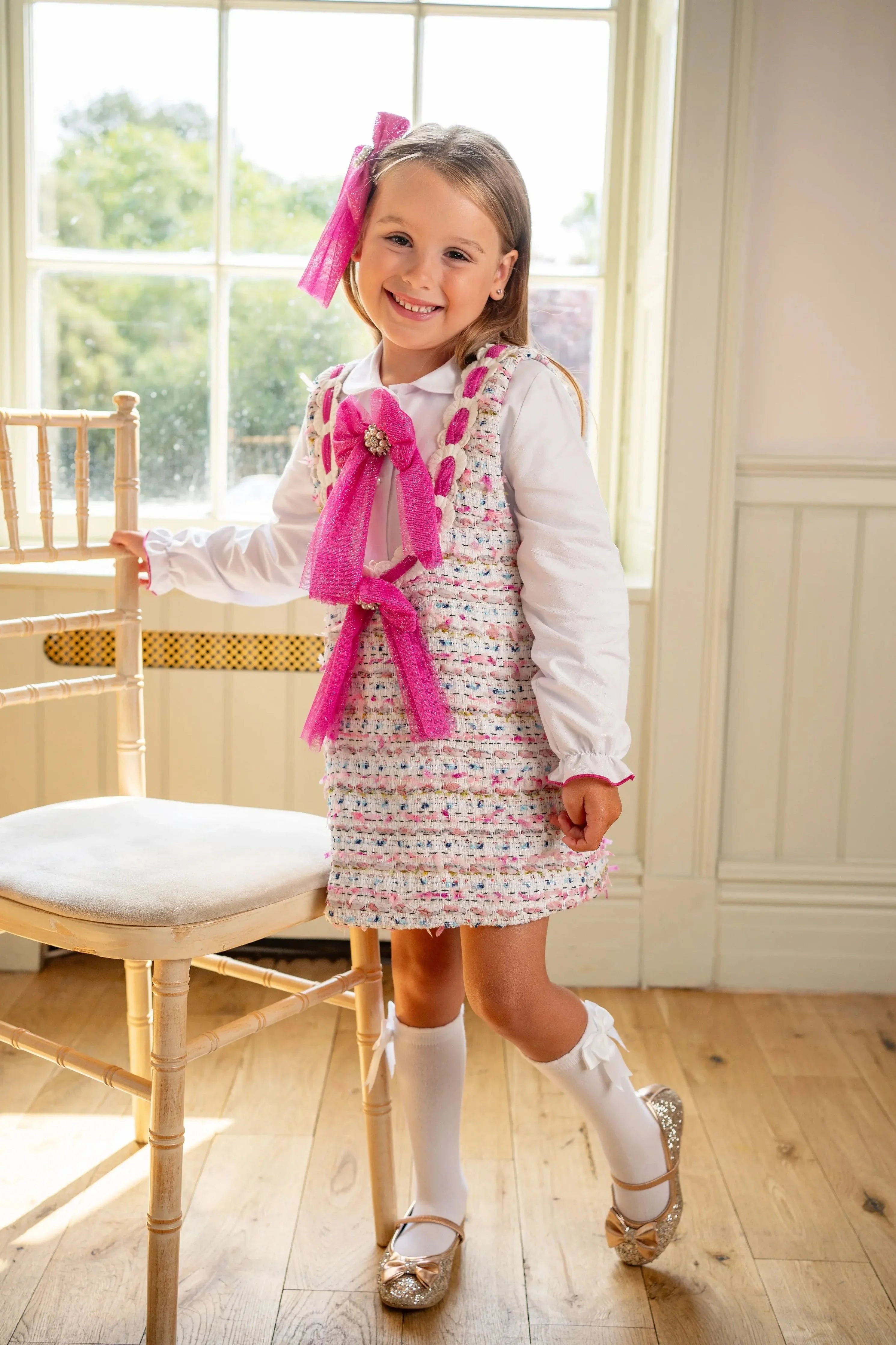 Smiling young girl in pink bow and tweed dress with white blouse, knee-high socks, and glitter shoes indoors