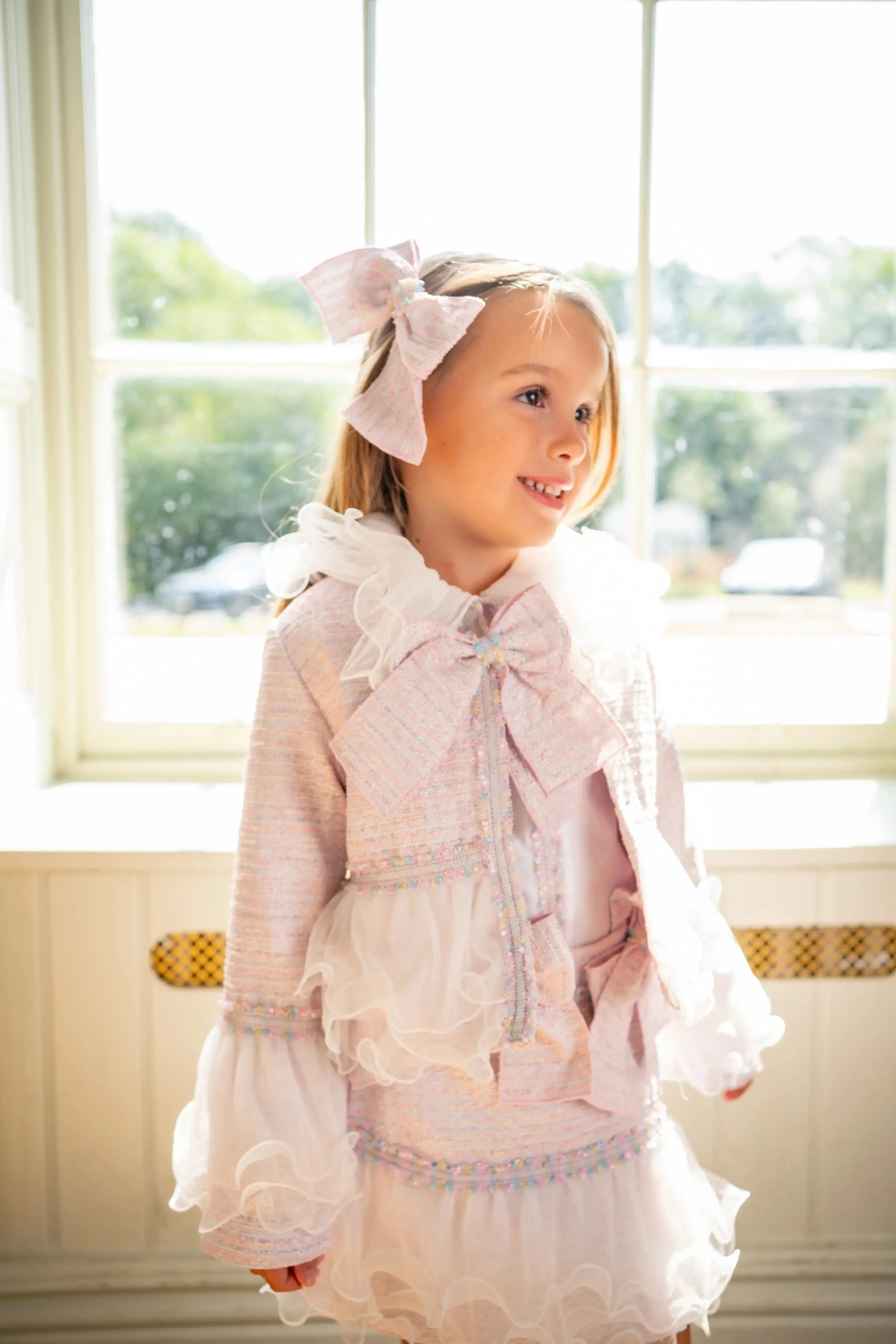 Smiling young girl in pastel pink bow and ruffled dress standing by window with natural light