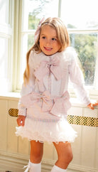 Smiling girl in white frilly dress with pink bows standing by bright window indoors