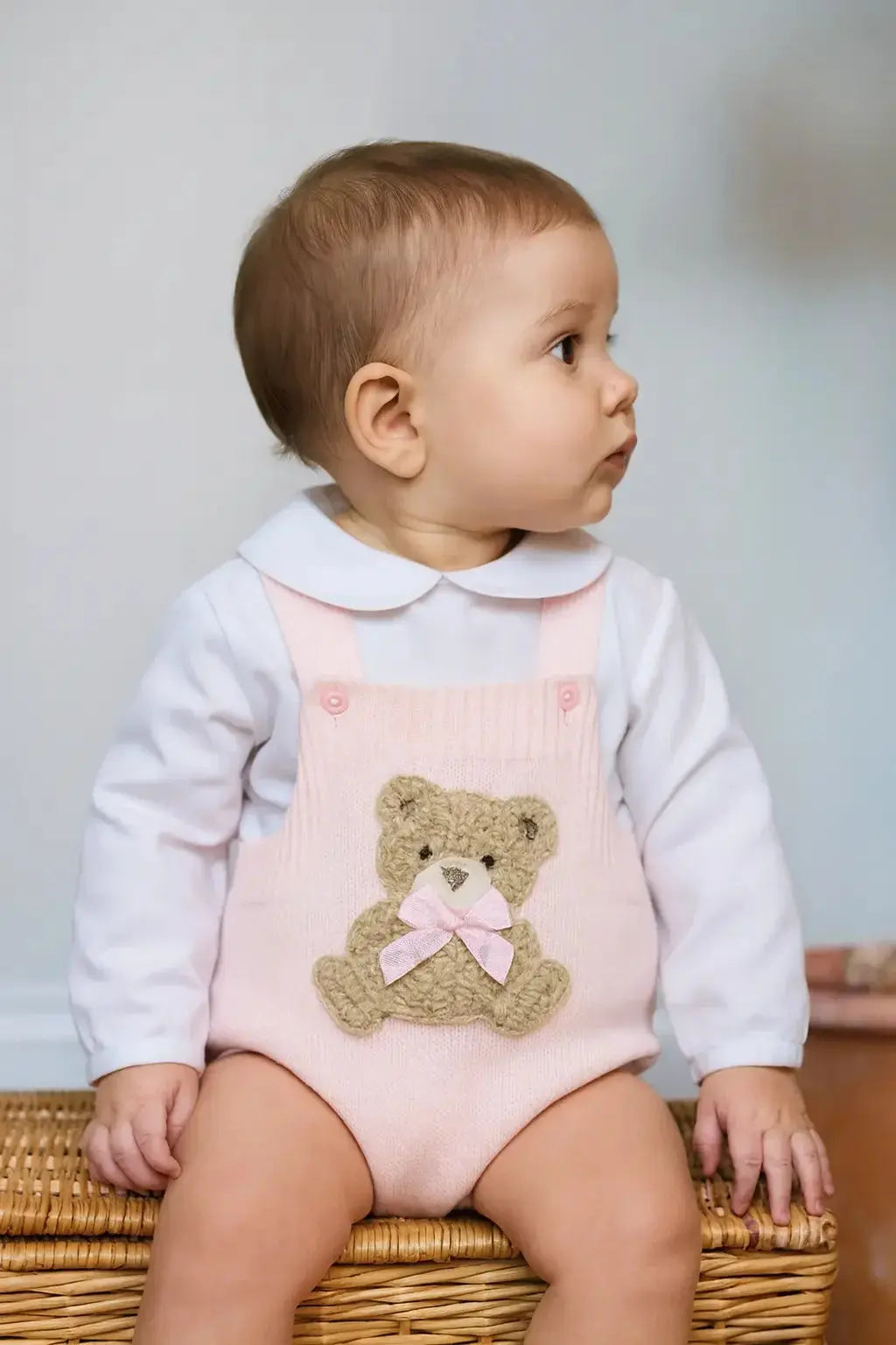 Baby wearing pink knitted romper with teddy bear and white shirt sitting on wicker basket