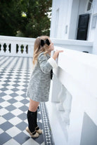 Young girl in black tweed dress and knee-high socks with bow, standing on black and white checkered floor balcony.