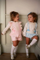 Two toddlers wearing pink and blue gingham pinafore outfits with white blouses and knee-high socks