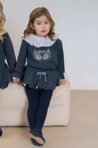 Young girl in navy swan print top with lace collar, matching skirt and tights, sitting on beige couch