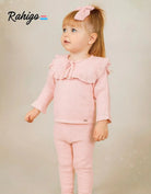 Toddler girl wearing a pink knitted tracksuit with ruffled collar and bow headband on beige background