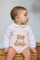 Baby wearing pink knitted romper with teddy bear and white shirt sitting on wicker basket
