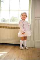 Smiling toddler girl in pink dress with ruffles, white knee-high socks, and gold shoes by window