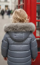 Child wearing gray puffer jacket with fur hood, standing near red British phone booth in city street