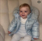 Smiling baby wearing light blue puffer jacket with fur hood sitting on beige textured armchair