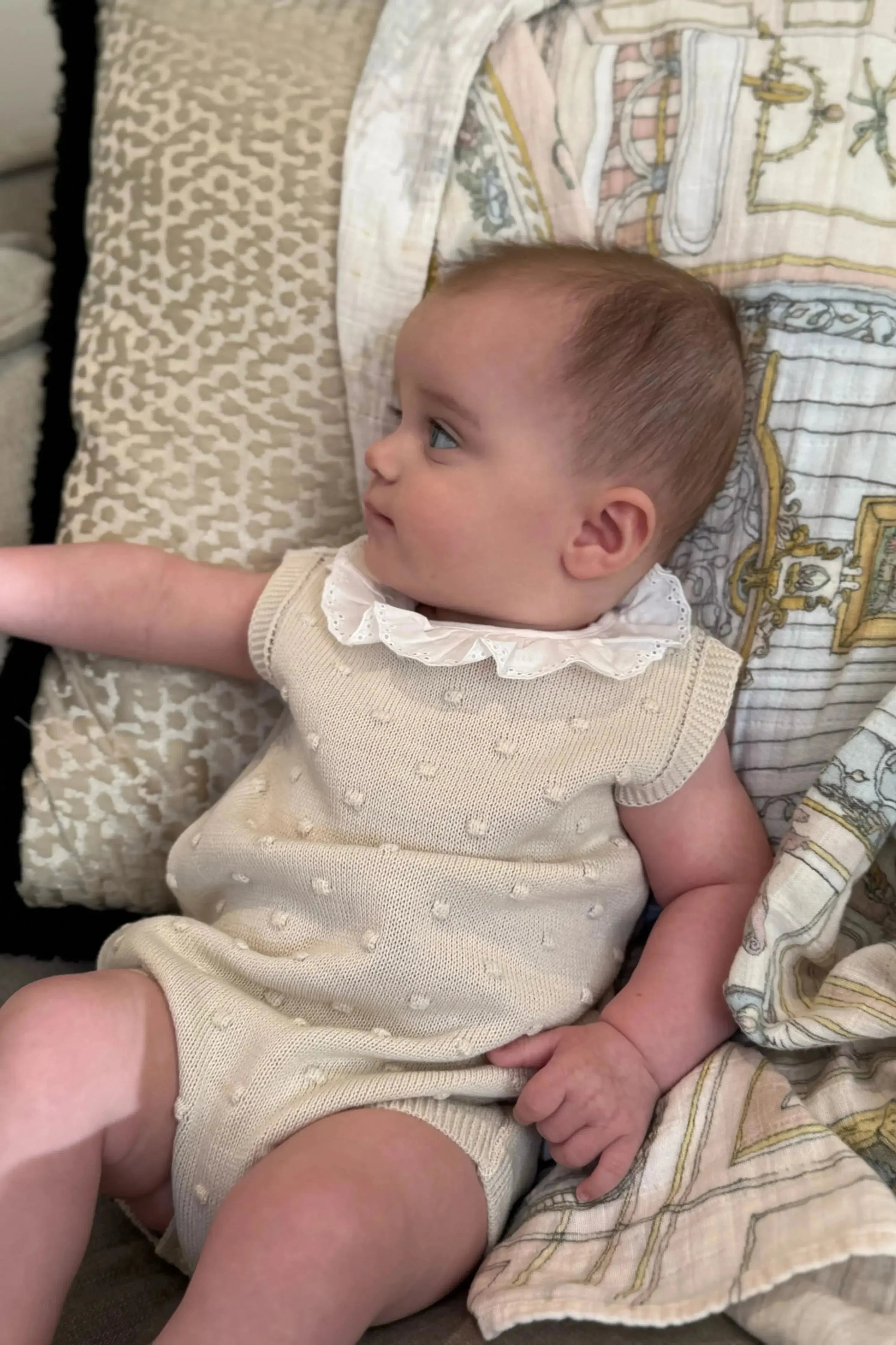 Baby girl wearing camel knitted frill romper lying on patterned cushions and blanket