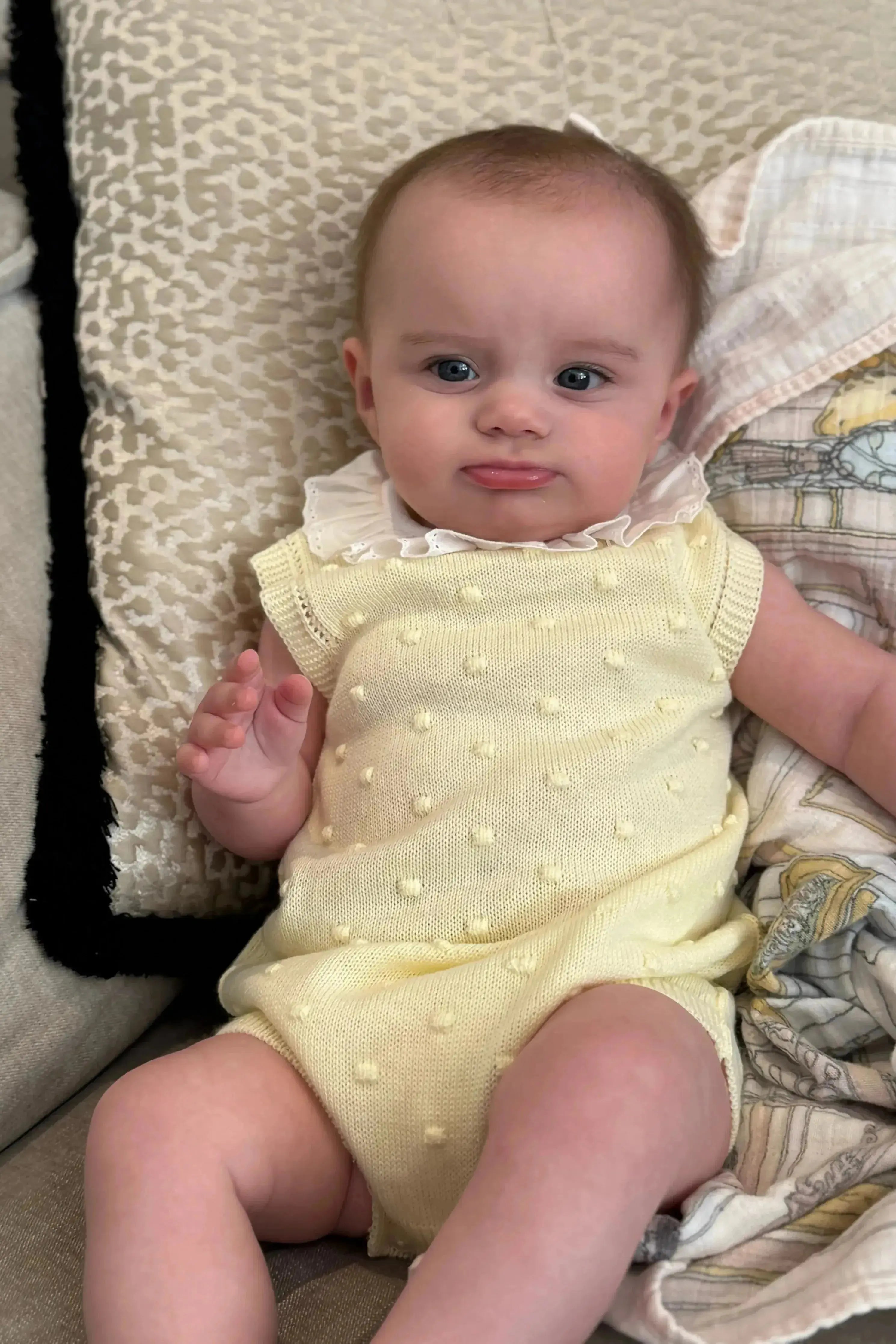 Baby in a yellow textured romper with a ruffled collar sitting on a patterned cushion and soft blanket