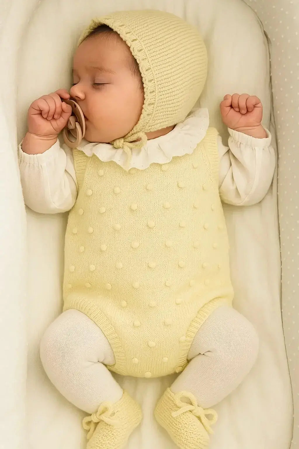 Sleeping baby in yellow knit bonnet and outfit with white tights and pacifier in soft crib