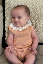 Baby girl smiling wearing peach knitted romper with white frill collar sitting on patterned cushion