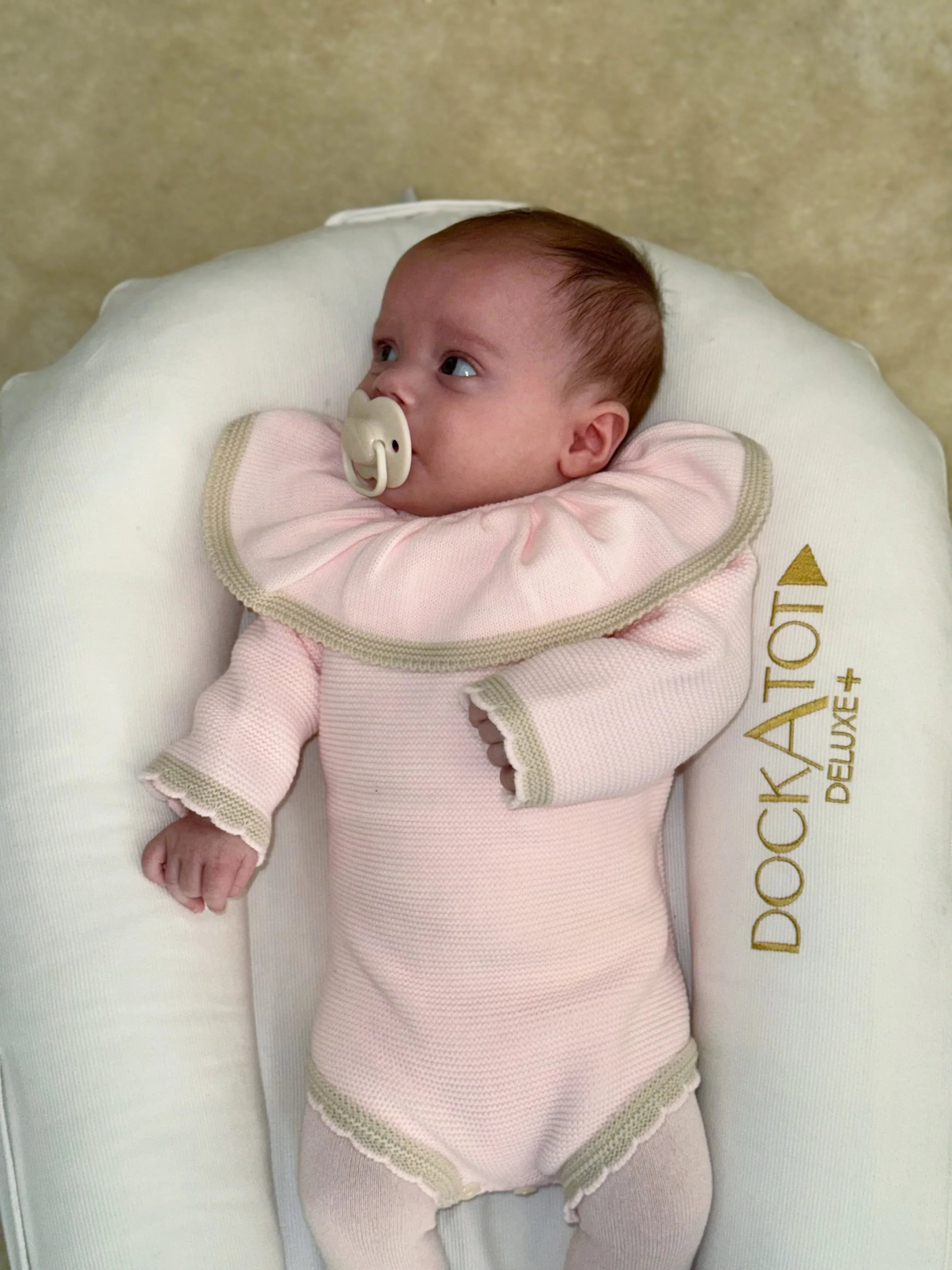 Baby in pink knit outfit with large collar lying in white DockATot Deluxe+ baby lounger