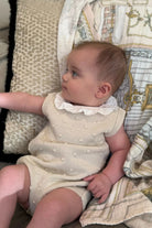 Baby girl wearing camel knitted frill romper lying on patterned cushions and blanket