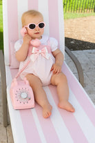 Baby girl wearing pink sunglasses and white pink summer romper sitting on striped lounge chair holding pink rotary phone