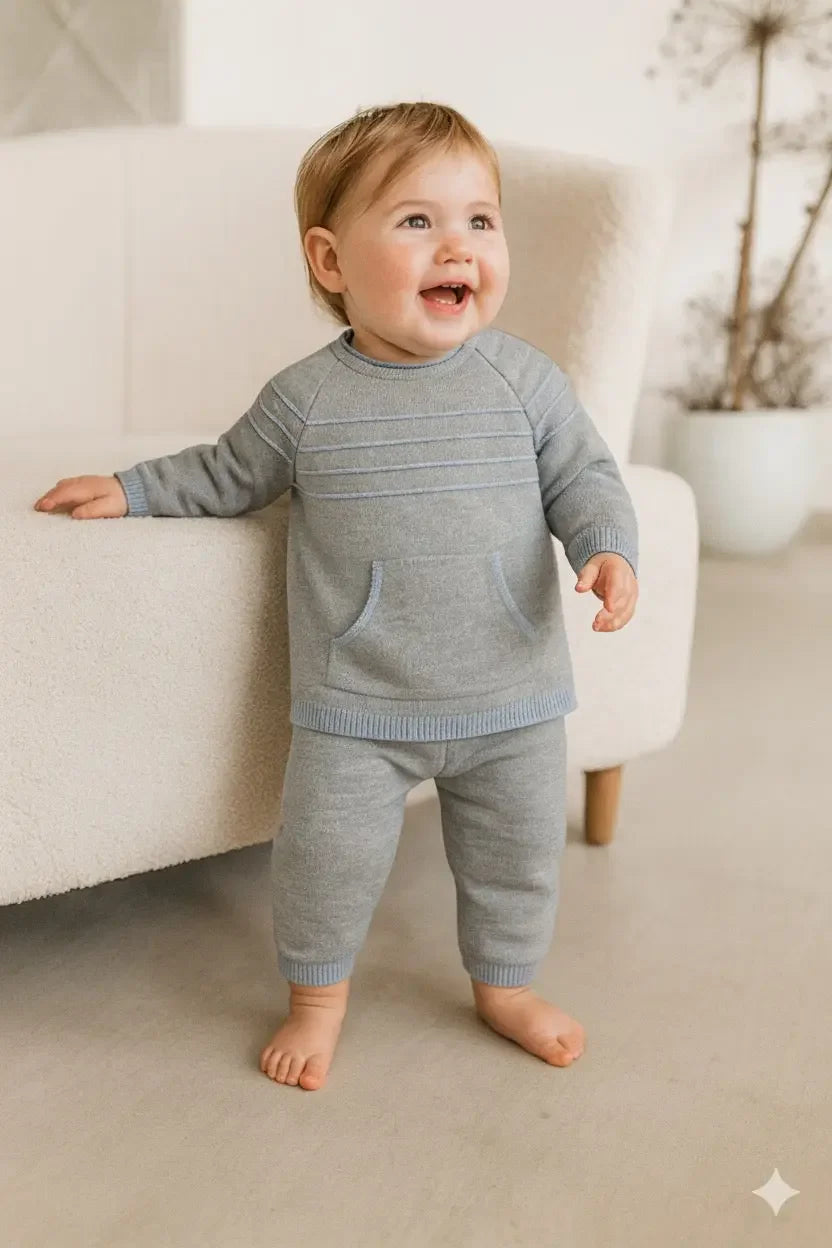 Smiling toddler standing barefoot in light gray knit sweater and pants indoors near beige armchair