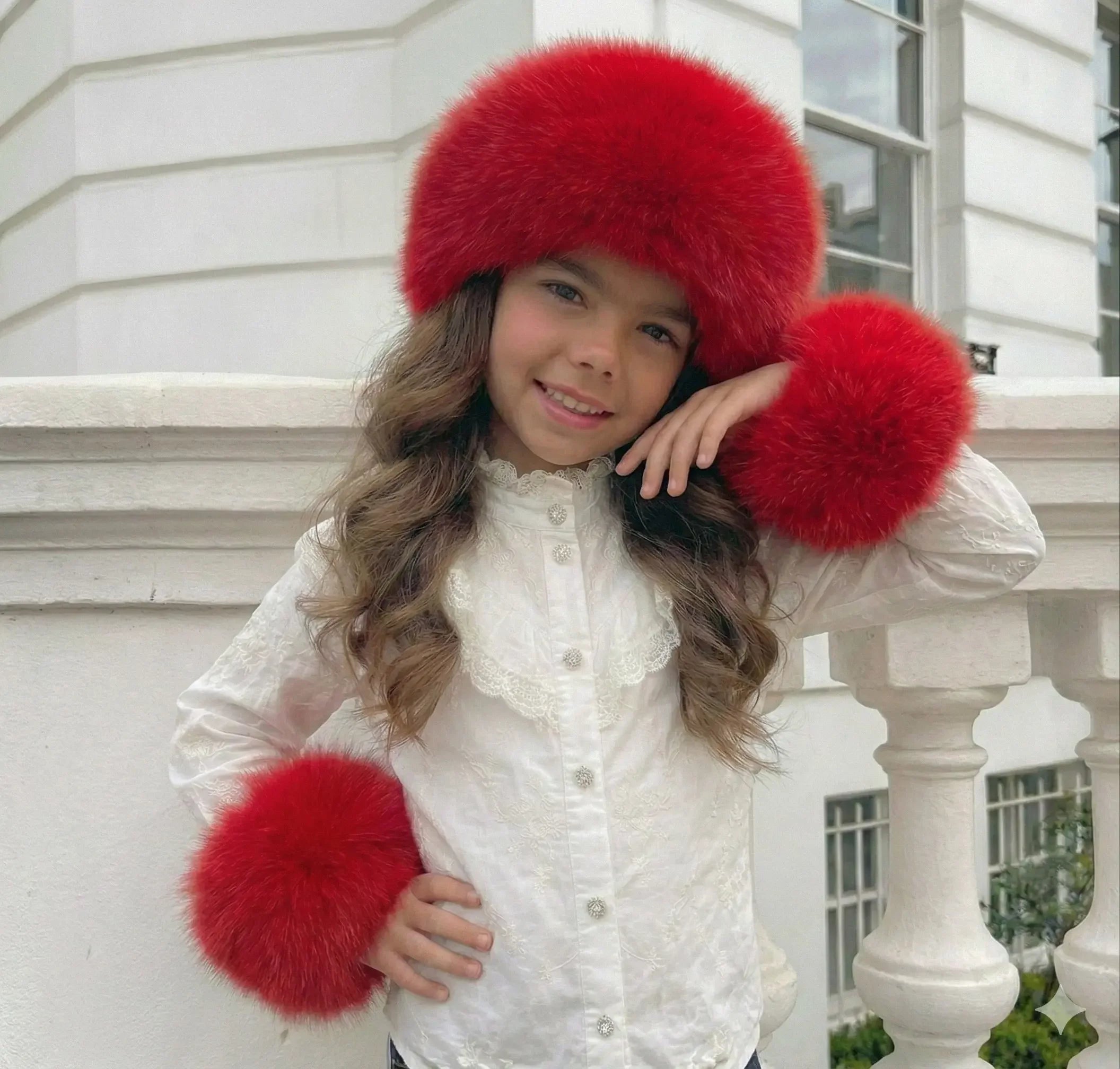 Smiling girl wearing white blouse with lace and red fluffy hat and cuffs against white building