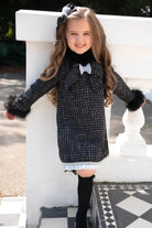Smiling young girl in black and white checkered dress with fur cuffs and bow, standing outdoors