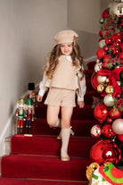 Girl in beige knitted shorts set and beret walking down red carpet stairs with Christmas decor and nutcracker figurines