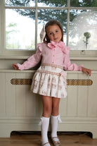 Young girl wearing a pink blouse with bow and a white patterned skirt standing indoors by a window