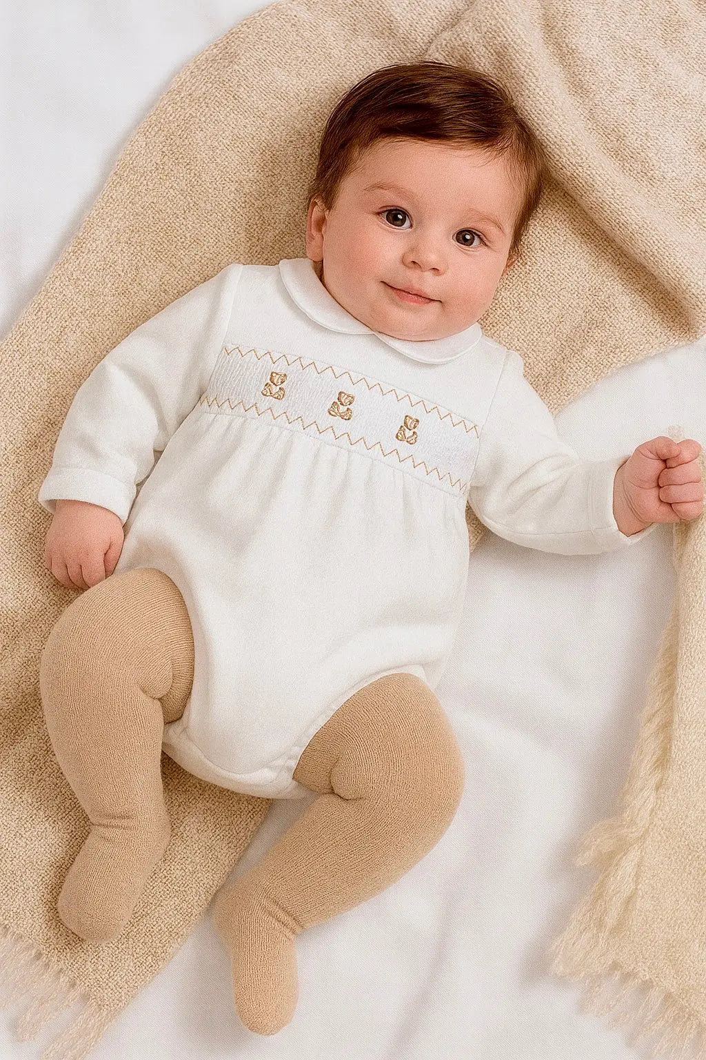 Baby wearing a white embroidered romper with beige tights lying on a textured blanket.