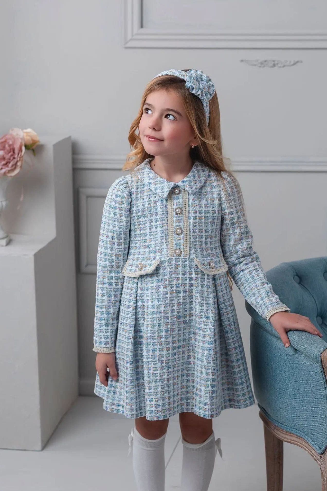 Young girl in a checkered dress with a headband, standing in a room with a decorative wall and chair.