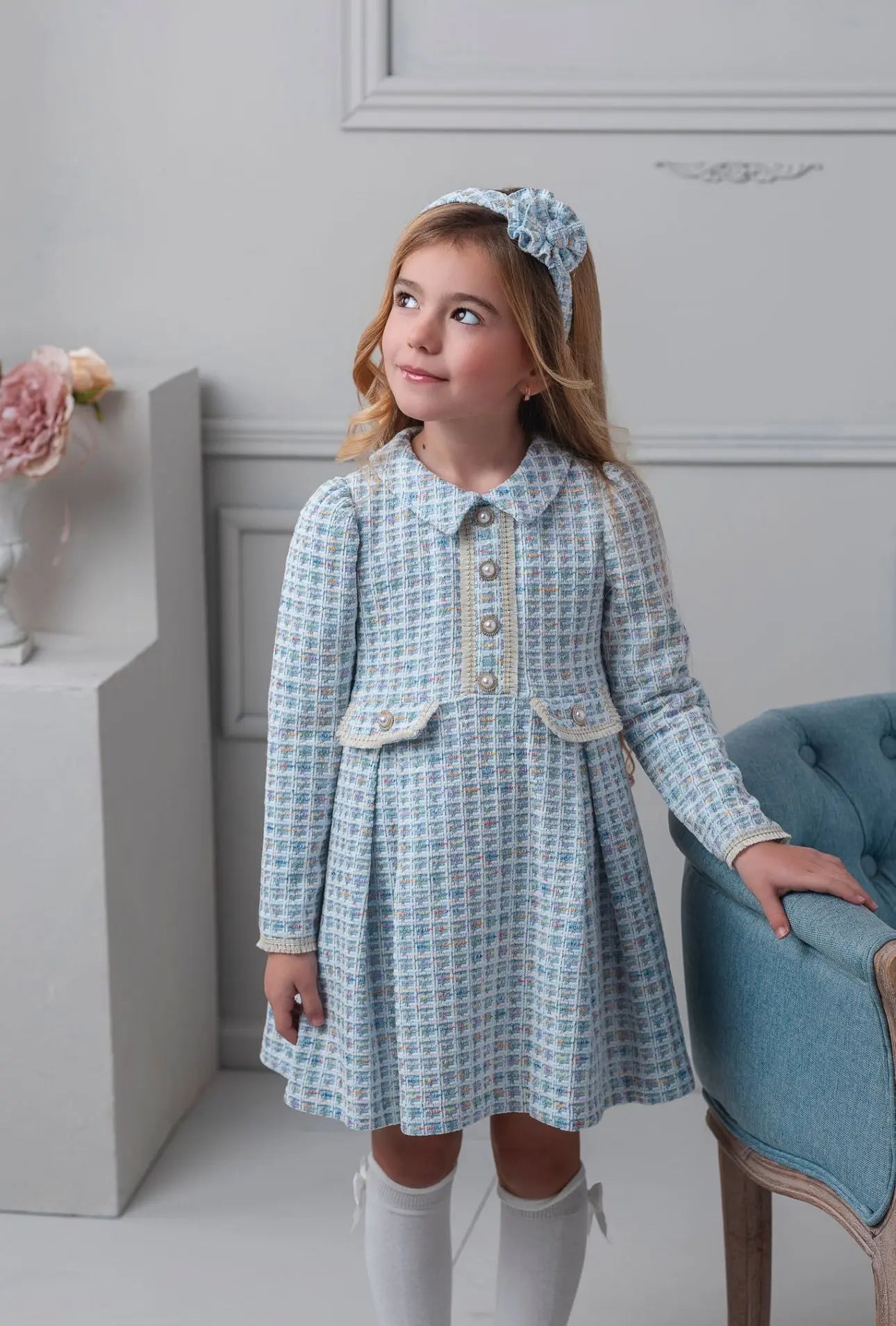 Young girl in a checkered dress with a headband, standing in a room with a decorative wall and chair.