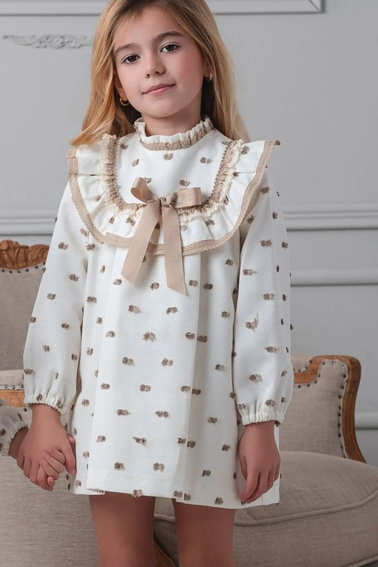 Young girl wearing a white dress with ruffled collar and bow in a room with a gray wall and wooden chair.