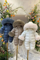 Three padded baby snowsuits with fur-lined hoods in navy, taupe, and white displayed on stands with floral backdrop