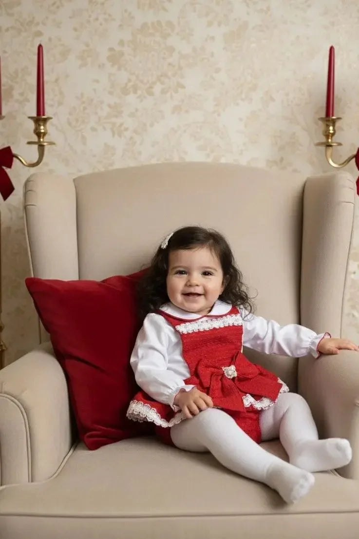 Smiling baby girl in red dress with white lace, sitting on beige armchair with red cushion