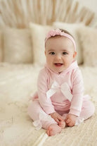 Smiling baby girl in pink outfit with bows and floral headband sitting on cream textured bed