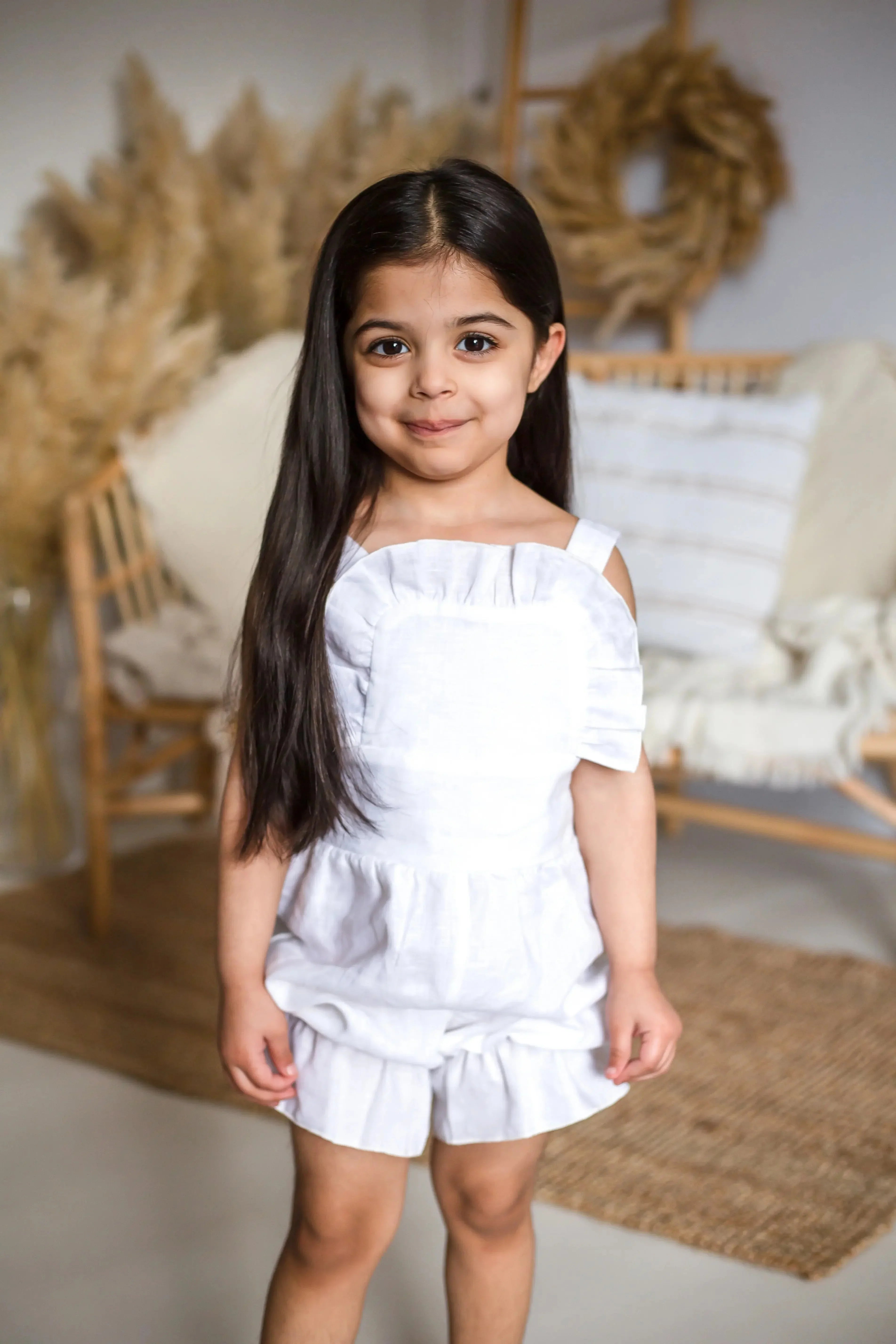 Young girl wearing an ivory linen frilly jumpsuit standing indoors with home decor background