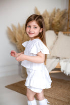 Smiling young girl in ivory linen pleated short set with ruffled sleeves in cozy neutral room