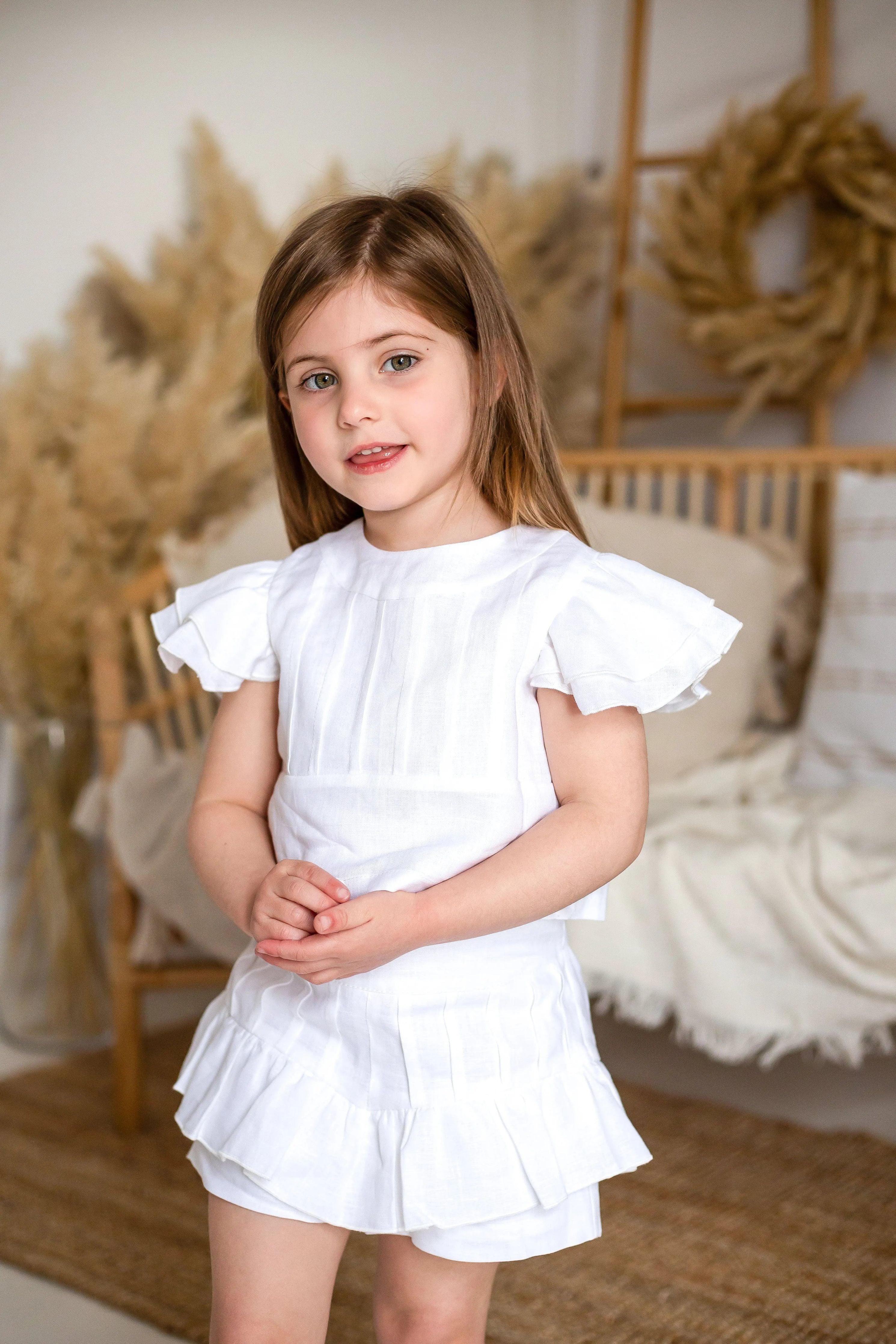 Young girl wearing an ivory pleated linen short set with ruffle sleeves in a neutral-toned room