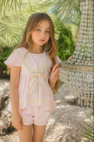 Young girl wearing a pink sailor short set with bow detail, standing outdoors by a crystal chandelier