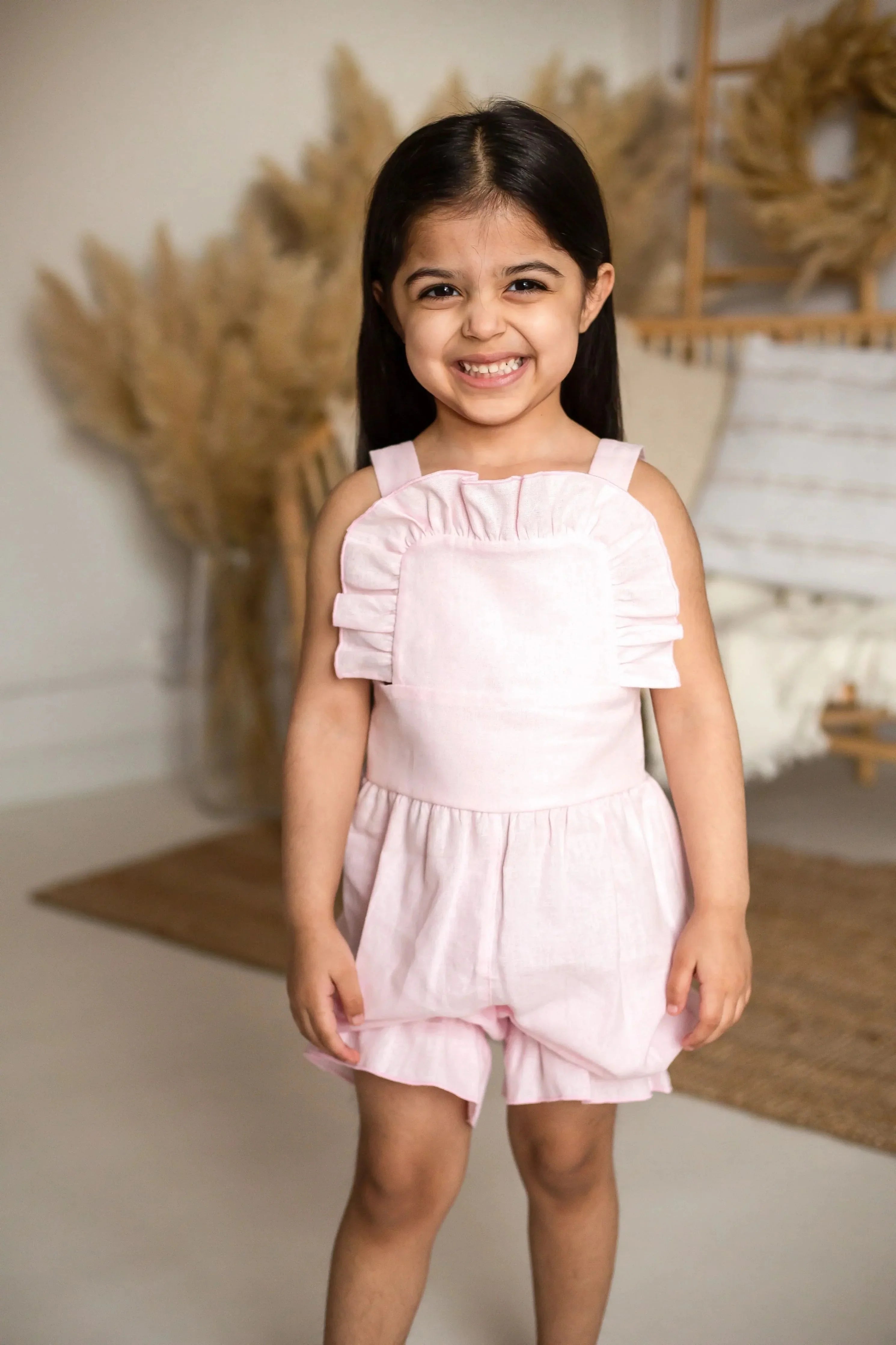 Smiling young girl wearing a pink linen frilly jumpsuit in a cozy, neutral-toned room
