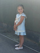 Young girl wearing baby blue linen sailor dress with cream bow and ruffled collar, standing outdoors