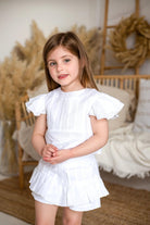 Young girl wearing an ivory pleated linen short set with ruffle sleeves in a neutral-toned room