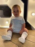 Baby boy sitting on wooden floor wearing white romper, knee-high socks, and white shoes under warm lighting