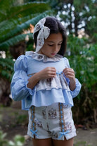 Girl wearing light blue and white outfit with carousel print shorts and matching hair bow in garden